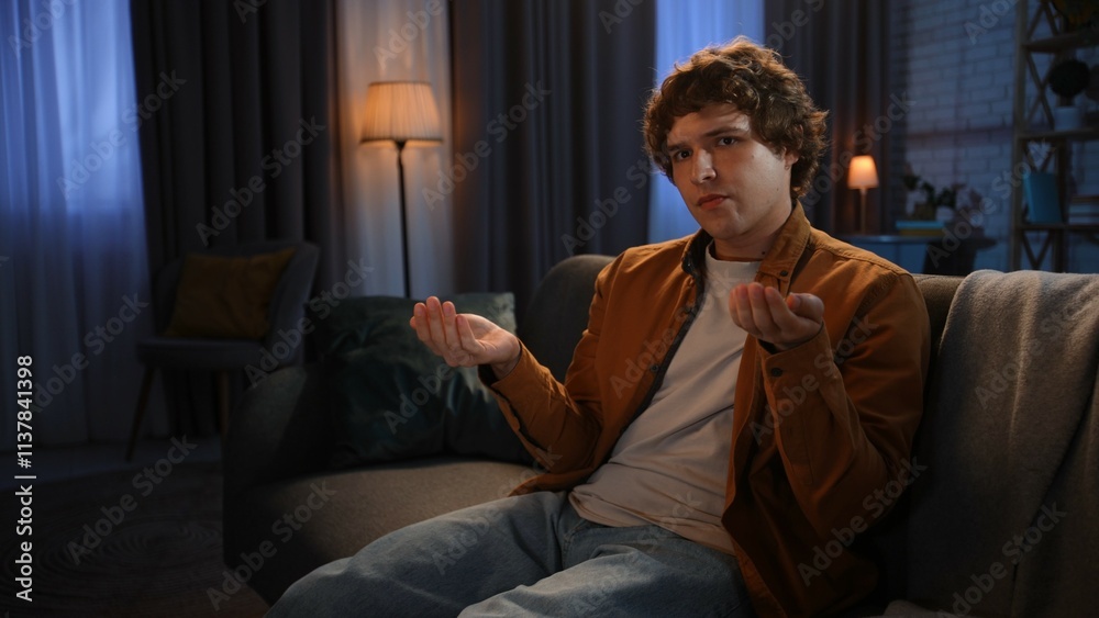Fototapeta premium Young man spending evening at home sitting on sofa in living room, holding coins in hands looking upset at camera, disappointed hopeless expression.