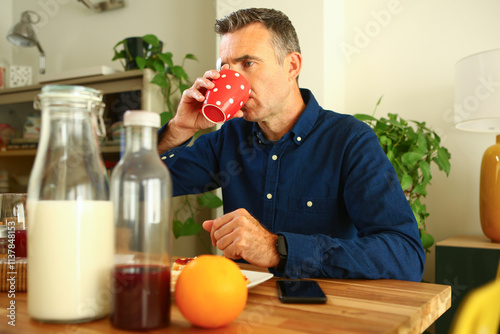 A 45 year old man dressed in a blue shirt drinking coffee from a red polka dot mug having breakfast before going to work photo