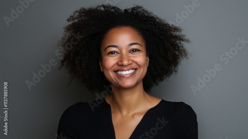 A poised and cheerful Black woman with curly natural hair, her smile radiating positivity in a minimalistic gray studio setting photo