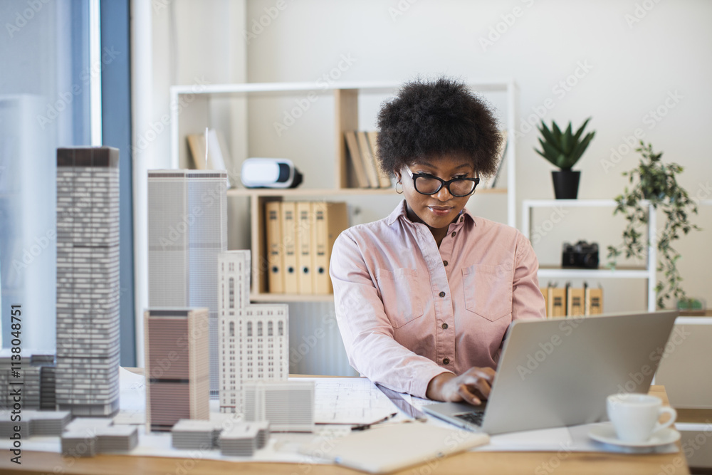 Fototapeta premium African woman architect working on laptop to design modern city model in office setting. Professional, focused, middle-aged woman with architectural expertise and creativity.