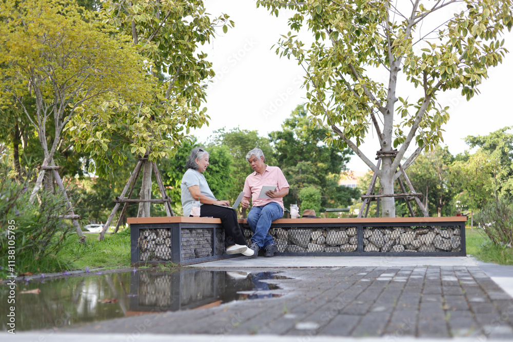 Fototapeta premium Senior couple sitting on a bench in park, Asian senior man and woman use laptop and digital tablet in park