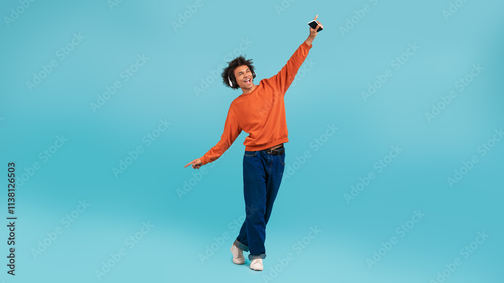 A young African American man with curly hair stands playfully against a vibrant light blue background. He raises one arm joyfully while holding a smartphone, expressing a carefree lifestyle. A young African American man with curly hair stands playfully against a vibrant light blue background. He raises one arm joyfully while holding a smartphone, expressing a carefree lifestyle.