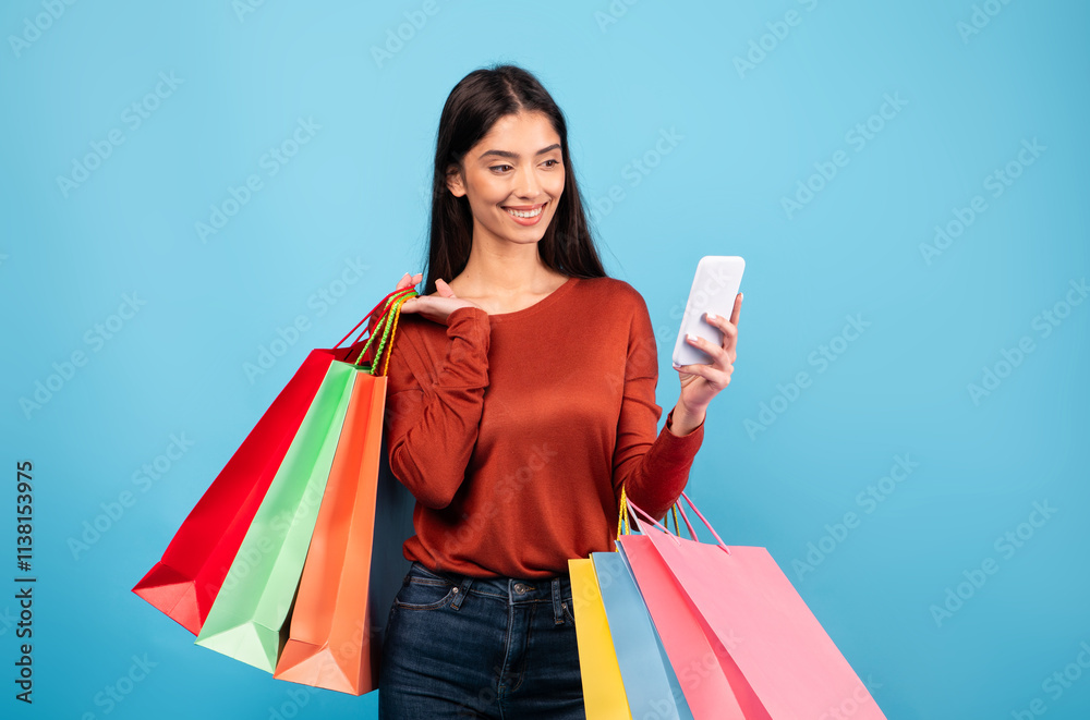 A millennial woman with brunette hair happily holds colorful shopping bags in one hand while looking at her smartphone with a smile. This lively atmosphere reflects her modern lifestyle.