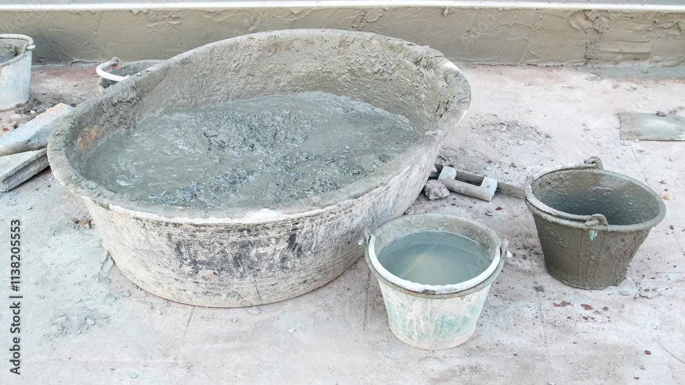 Close-up view of cement mixing tools at a construction site, featuring a large basin filled with mixed cement, two smaller buckets and surrounding concrete work surfaces. Close-up view of cement mixing tools at a construction site, featuring a large basin filled with mixed cement, two smaller buckets and surrounding concrete work surfaces.