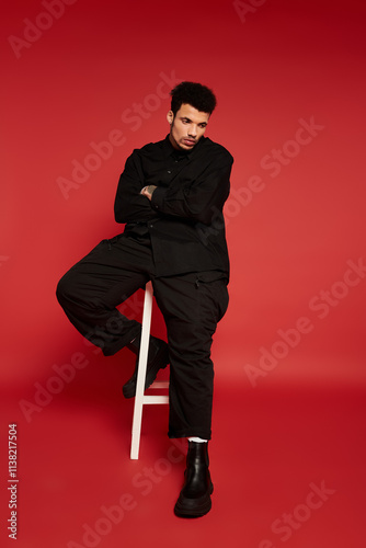 A young handsome man showcases a stylish outfit while seated on a stool against a red backdrop. photo