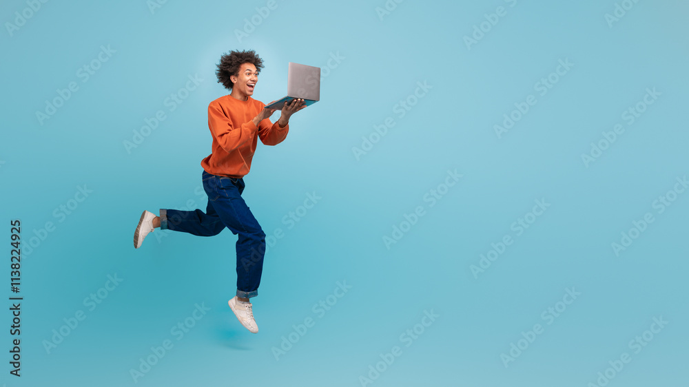 A young African American man with curly hair is leaping with excitement while holding a laptop. He conveys a sense of enthusiasm and energy in a colorful space, copy space A young African American man with curly hair is leaping with excitement while holding a laptop. He conveys a sense of enthusiasm and energy in a colorful space, copy space