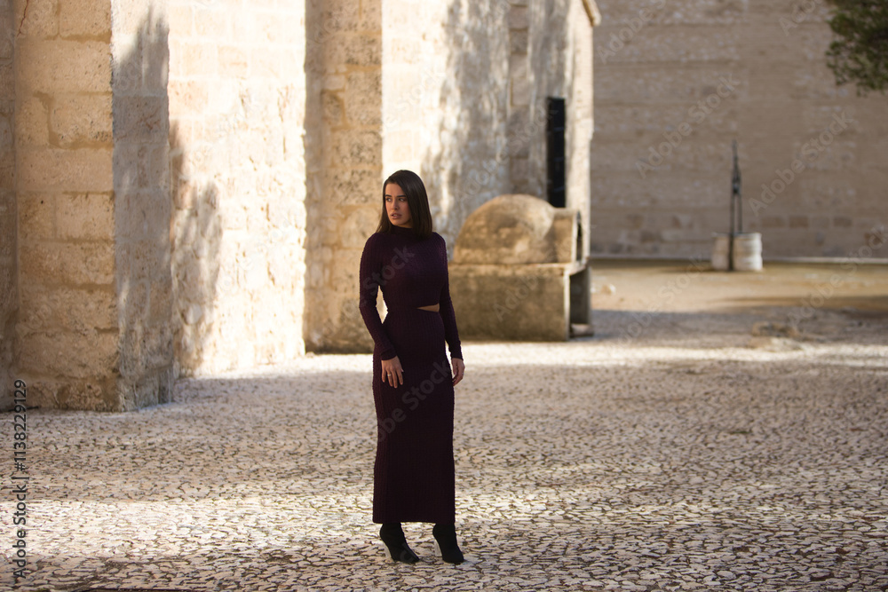 pretty young woman dressed in elegant violet dress posing on a cobblestone street next to a monastery in a village in the city of Seville, Spain. The woman makes different expressions and gestures. pretty young woman dressed in elegant violet dress posing on a cobblestone street next to a monastery in a village in the city of Seville, Spain. The woman makes different expressions and gestures.