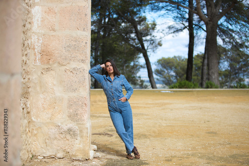 Young and beautiful Latin woman, brunette with short hair, dressed in a denim jumpsuit leaning sideways on a stone wall of an old monastery in the province of Seville, Spain.