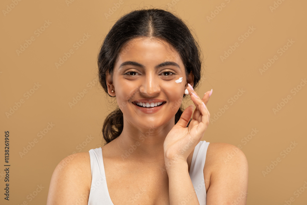 A young Indian woman with beautiful skin applies a skincare product while smiling. She is pampering herself in an isolated space that exudes calmness and relaxation. A young Indian woman with beautiful skin applies a skincare product while smiling. She is pampering herself in an isolated space that exudes calmness and relaxation.