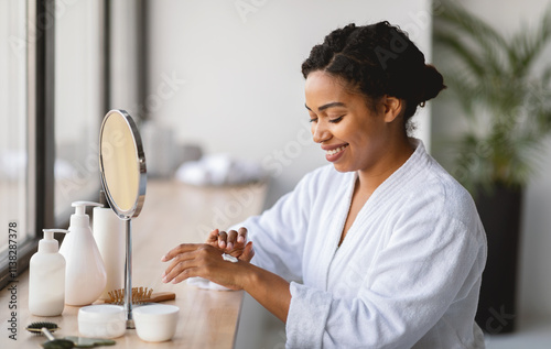 African American woman in a bathrobe is applying skincare products at a vanity. Natural light fills the room, creating a serene ambiance. photo