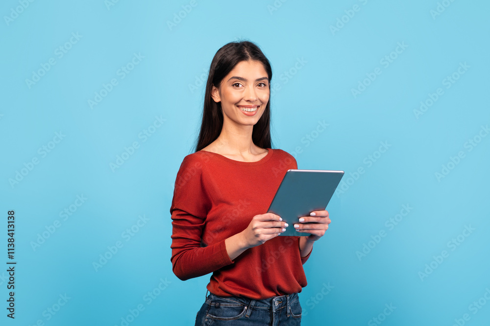 A millennial woman with long brunette hair wears a red sweater and smiles while holding a tablet. The bright blue background enhances her cheerful demeanor, creating a lively atmosphere. A millennial woman with long brunette hair wears a red sweater and smiles while holding a tablet. The bright blue background enhances her cheerful demeanor, creating a lively atmosphere.