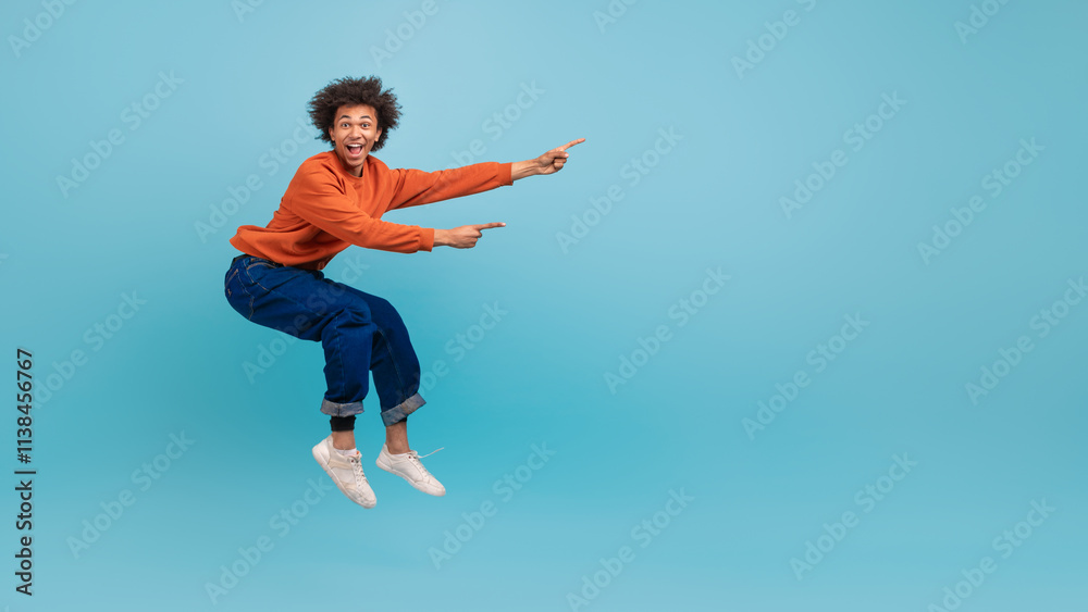A young African American man with curly hair is jumping in excitement against a bright blue background. He is wearing an orange sweater and denim jeans, creating a lively atmosphere, copy space A young African American man with curly hair is jumping in excitement against a bright blue background. He is wearing an orange sweater and denim jeans, creating a lively atmosphere, copy space