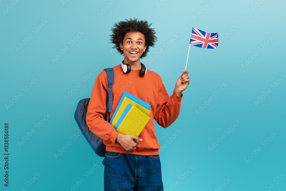 A young African American man with curly hair smiles brightly while holding a small British flag. He wears a casual orange sweatshirt and headphones, carrying yellow and blue folders. A young African American man with curly hair smiles brightly while holding a small British flag. He wears a casual orange sweatshirt and headphones, carrying yellow and blue folders.