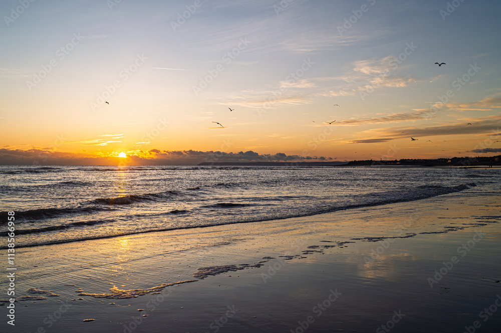 West St Leonards beach at sunset. The scene shows the beach at low ride, exposing the sand. Waves roll into the beach. The setting sun creates contrasting colours and deep shadows. West St Leonards beach at sunset. The scene shows the beach at low ride, exposing the sand. Waves roll into the beach. The setting sun creates contrasting colours and deep shadows.