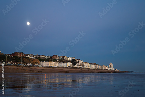 West St Leonards at Dusk. The scene shows the seafront buildings reflected in the wet sand. The tide is out and the scene is shot at dusk. The moon rises in the background.