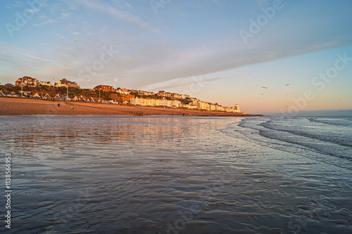 Fototapeta Naklejka Na Ścianę i Meble -  West St Leonards at Dusk. The scene shows the seafront buildings reflected in the wet sand. The tide is out and the scene is shot at dusk. The moon rises in the background.