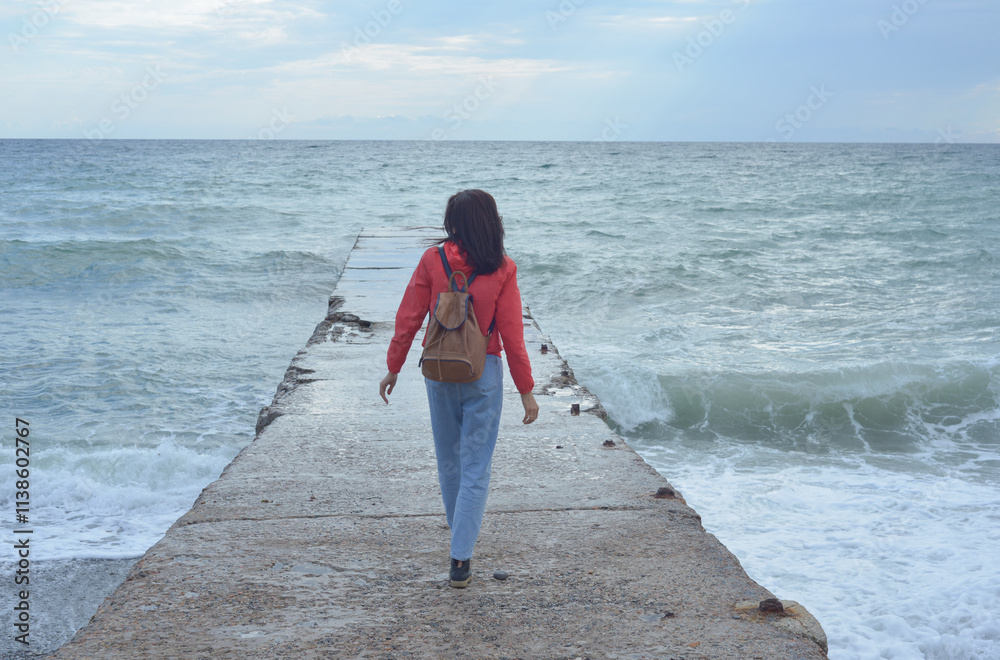 Rear view of a young woman tourist with a backpack standing on the pier and waves hitting the pier in a storm. City of Sudak, Crimea.