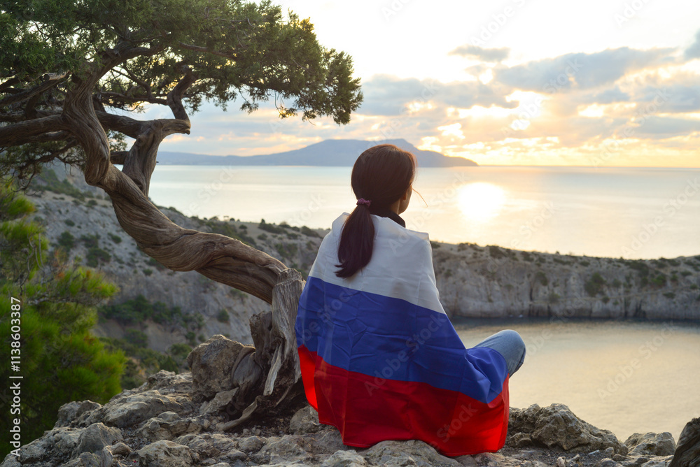 Young traveler with the Russian flag on her shoulders on the top of Karaul-Oba mountain, Novy Svet village, Crimea, enjoying the sea view. A patriot with a tricolor travels through the mountains.