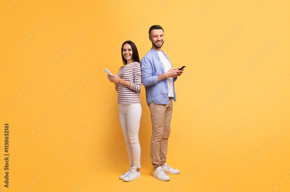 A couple stands back to back, smiling while using their smartphones. The cheerful atmosphere, highlighted by a vibrant yellow backdrop, captures their playful affection and connection. A couple stands back to back, smiling while using their smartphones. The cheerful atmosphere, highlighted by a vibrant yellow backdrop, captures their playful affection and connection.
