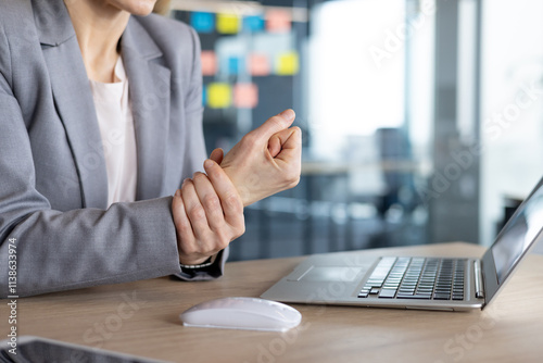 Fotografija Mature businesswoman seated at an office desk holds her wrist in discomfort, indicating potential repetitive strain injury likely caused by prolonged computer use