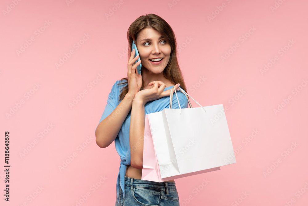 Studio portrait of casually dressed attractive woman holding shopping bags and looking aside with coquette face expression while accepting call on her smartphone, isolated on pastel pink background Studio portrait of casually dressed attractive woman holding shopping bags and looking aside with coquette face expression while accepting call on her smartphone, isolated on pastel pink background