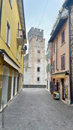 A narrow street with a tall white building in the background. The street is lined with shops and has a cobblestone surface