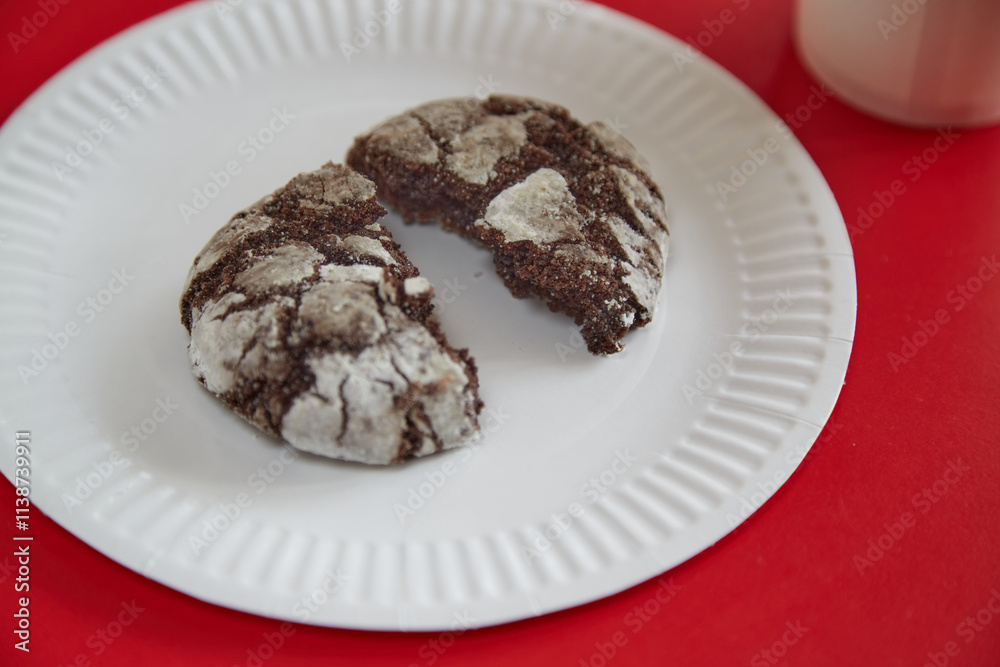 A broken chocolate cookie with cracks lying on a white paper plate against a red background, closeup A broken chocolate cookie with cracks lying on a white paper plate against a red background, closeup