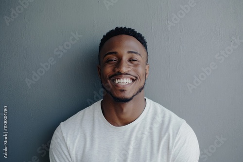 A cheerful man beams with a radiant smile, standing in front of a textured gray wall that adds warmth to the cozy indoor environment