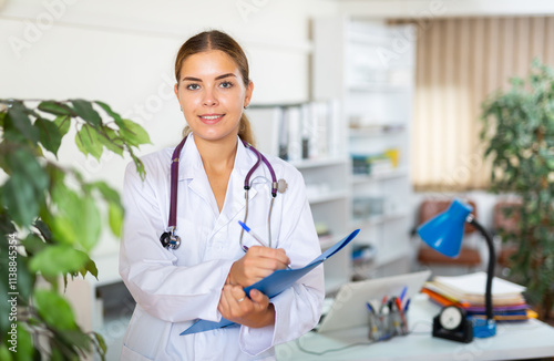 Young female doctor in white gown and stethoscope on shoulders standing in hall of clinic. She's holding folder and pan in hands. photo