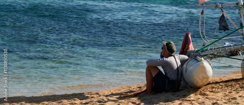 A lone man in a sailor's shirt sits by a sailing catamaran on an offshore sandy beach at the water's edge. He stares out at the sea in the distance. Copy space. photo