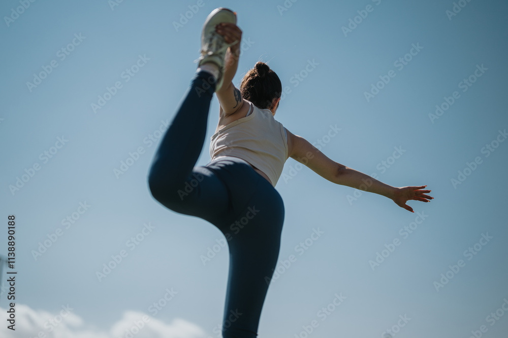 Obraz premium A woman performing a yoga pose outdoors, balancing gracefully under a clear blue sky. Her athletic form and tranquil environment embody fitness and mindfulness in nature.