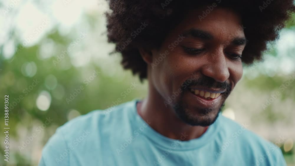 Closeup portrait of young African American man in light blue t-shirt sitting on city park bench and looks at photos, videos in his mobile phone. Camera moving forwards approaching. Closeup portrait of young African American man in light blue t-shirt sitting on city park bench and looks at photos, videos in his mobile phone. Camera moving forwards approaching.