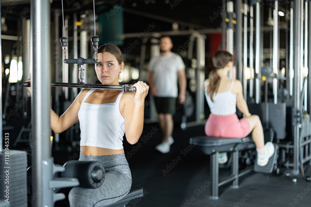 Active athlete young woman during exercises on gym apparatus for training muscles of hands tris, in sport club. Visitor using cable crossover machine with some weightening substance Active athlete young woman during exercises on gym apparatus for training muscles of hands tris, in sport club. Visitor using cable crossover machine with some weightening substance