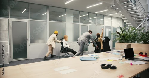 Cheerful men employees push businesswomen on office chairs