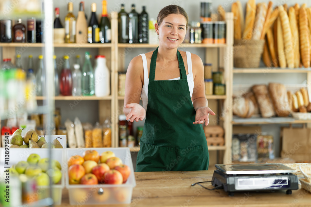 Obraz premium Portrait of positive female salesperson in apron behind supermarket counter
