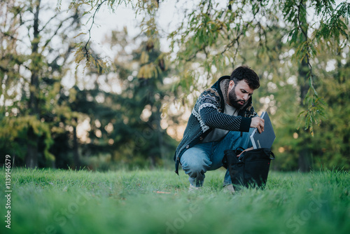 A man kneels on grass, packing a laptop into a backpack under lush green trees. The peaceful park environment suggests a productive and tranquil outdoor work setting.