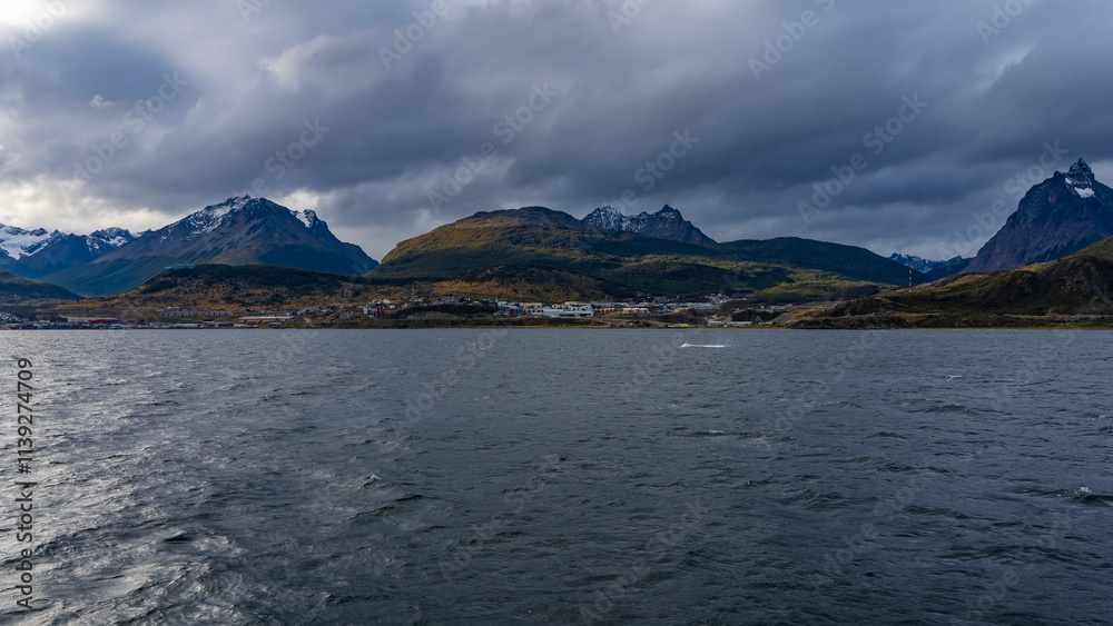 A beautiful mountain range with snow-capped peaks against a cloudy sky. A city at the foot of green hills, on the ocean. The surface of the Beagle Canal is in the foreground. Argentina. Patagonia. A beautiful mountain range with snow-capped peaks against a cloudy sky. A city at the foot of green hills, on the ocean. The surface of the Beagle Canal is in the foreground. Argentina. Patagonia.