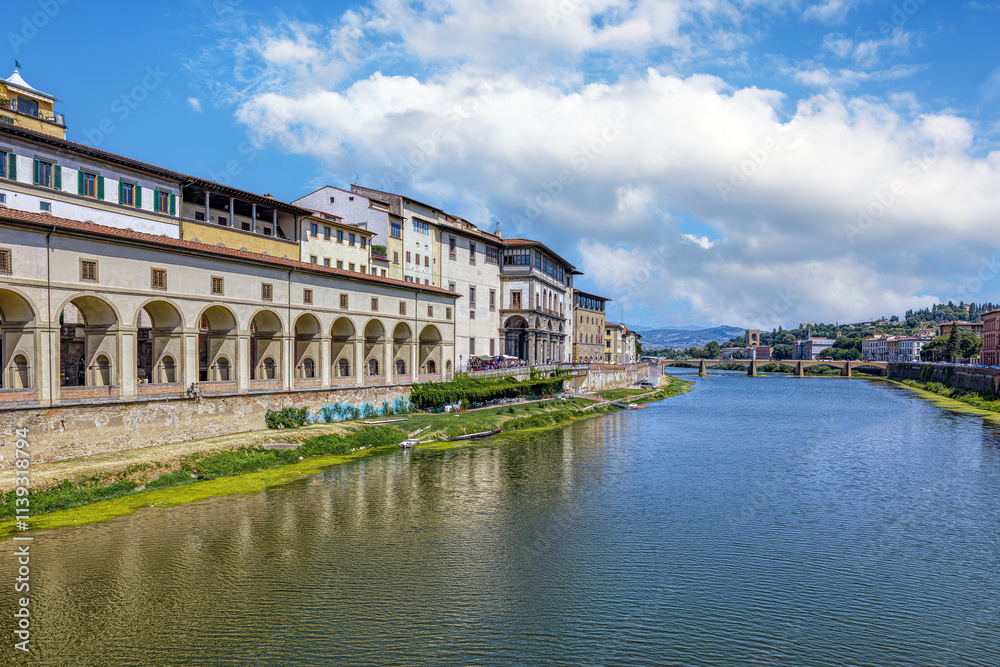 Colorful medieval houses sit above the Arno River in Florence, showcasing vibrant facades and historical architecture under blue cloudy sky.