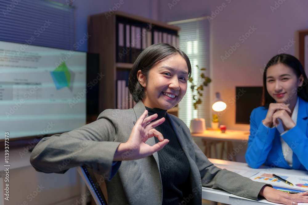 Two businesswomen working late in the office, reviewing charts and graphs on a computer screen, with one gesturing an ok sign Two businesswomen working late in the office, reviewing charts and graphs on a computer screen, with one gesturing an ok sign