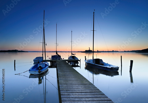 Wooden Pier by calm lake with boats before sunrise at dawn