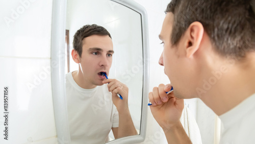 Young man brushing teeth with toothbrush while standing before mirror at home in the bathroom