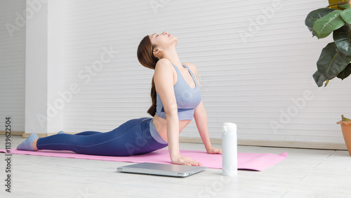 Asian young beautiful attractive woman wearing sportswear stretches body upward facing on mat, yoga workout pilates exercise training with laptop and water bottles in living room healthy wellness.