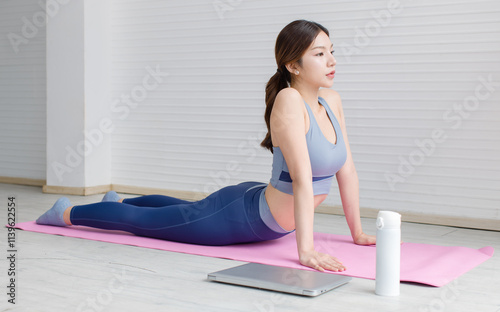 Asian young beautiful attractive woman wearing sportswear stretches body upward facing on mat, yoga workout pilates exercise training with laptop and water bottles in living room healthy wellness.