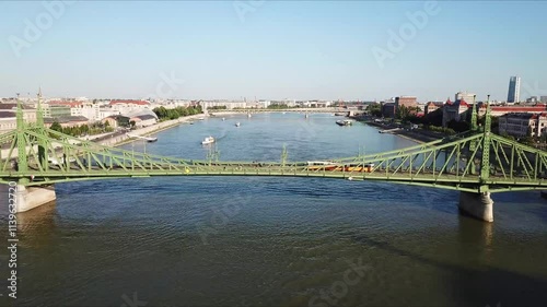 Liberty bridge, a green steel structure spanning the Danube river in Budapest, Hungary, features a tram crossing while boats navigate the waterway on a sunny day, drone fly over