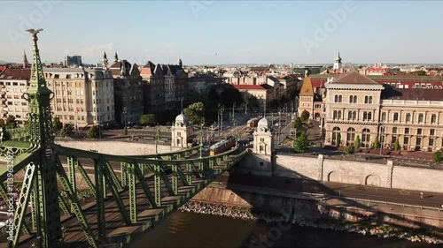 Aerial view of Liberty bridge, a green steel bridge over Danube river, with a tram and cars passing and Budapest cityscape in the background during a sunny day, Hungary, drone reveal shot