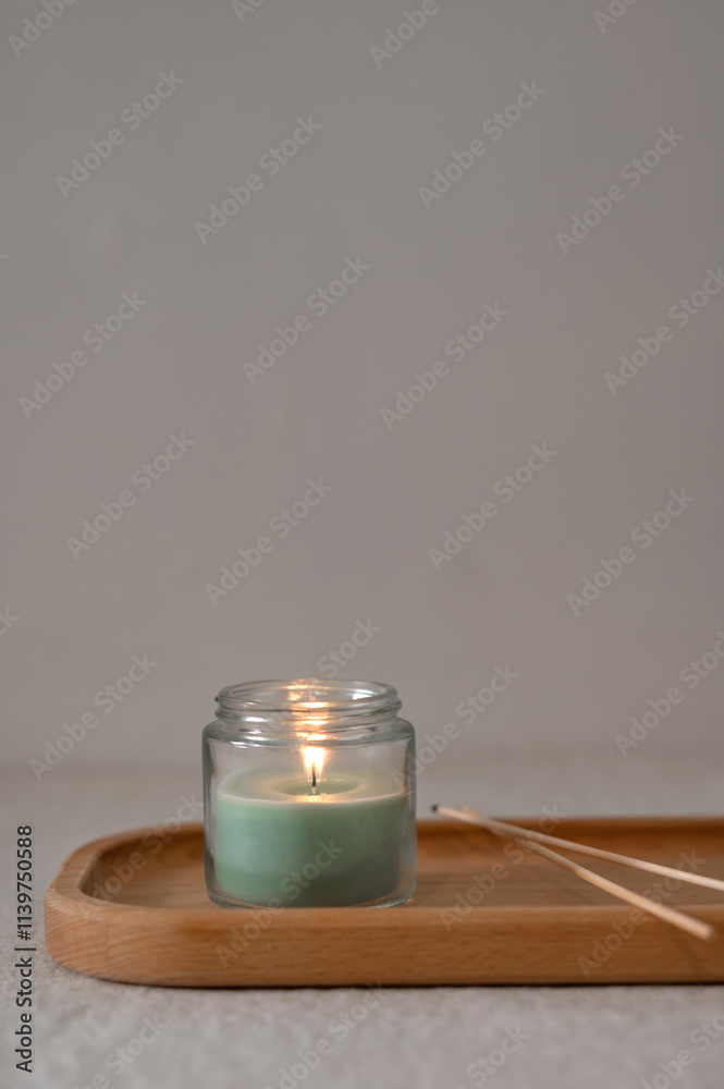 Scented green candle with soft dim light on wooden tray on table, empty neutral taupe gray beige wall background, cozy home aesthetic still life Scented green candle with soft dim light on wooden tray on table, empty neutral taupe gray beige wall background, cozy home aesthetic still life