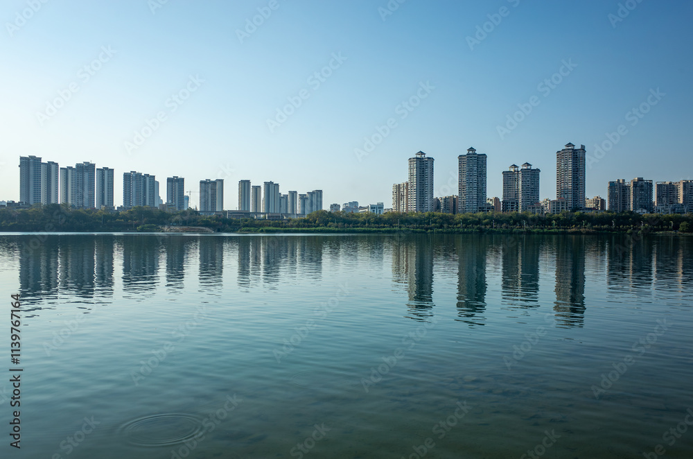 Naklejka premium Waterfront cityscape in Liuzhou, Guangxi, China. Calm river reflecting the skyline of modern residential buildings along the riverbank.Concept of urban development the issue of apartment oversupply.