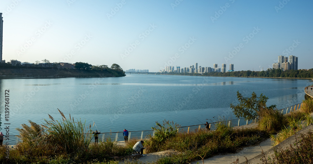A scenic panoramic view of a waterfront recreational walkway where people enjoy leisure activities against the backdrop of a modern city skyline. Liuzhou, China. A scenic panoramic view of a waterfront recreational walkway where people enjoy leisure activities against the backdrop of a modern city skyline. Liuzhou, China.