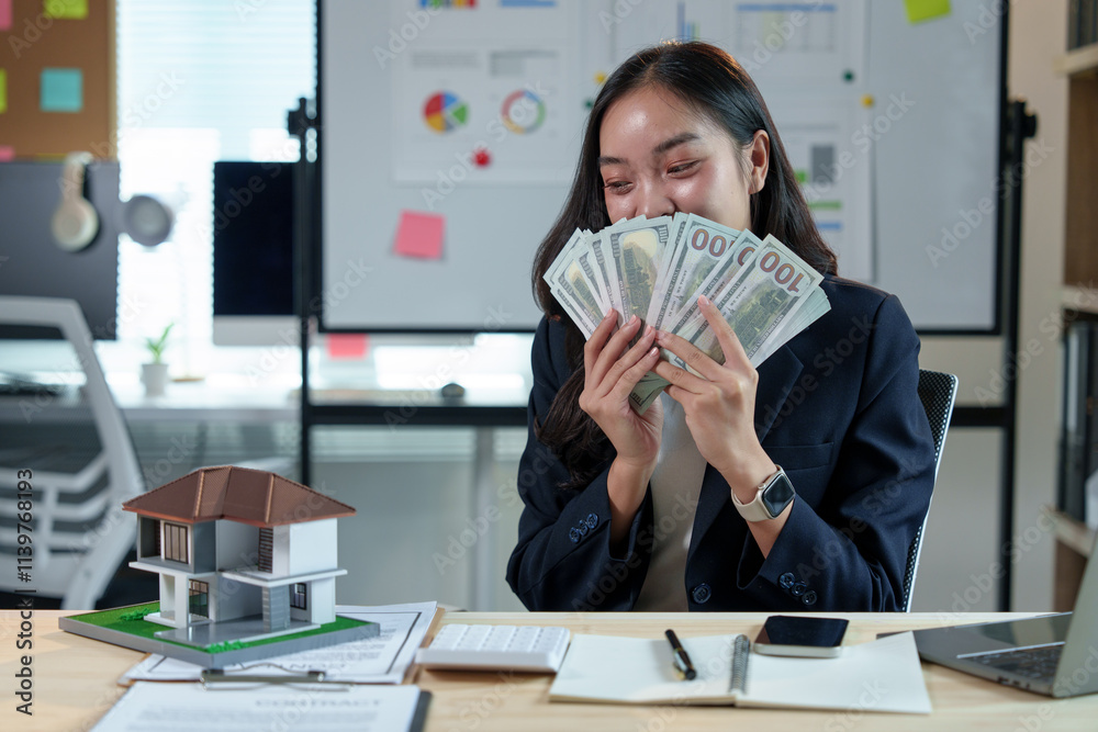 Asian sales girl, real estate agent holding money, counting money from house sale, customer's lease agreement, insurance while sitting at desk in office with model house in real estate project. Asian sales girl, real estate agent holding money, counting money from house sale, customer's lease agreement, insurance while sitting at desk in office with model house in real estate project.