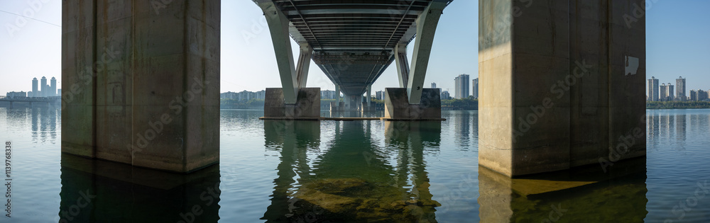 Panoramic view of underside of a bridge or pier structure reflected in the calm, still water below, city skyline in the distance. A sense of emptiness, and soothing, contemplative atmosphere. Panoramic view of underside of a bridge or pier structure reflected in the calm, still water below, city skyline in the distance. A sense of emptiness, and soothing, contemplative atmosphere.